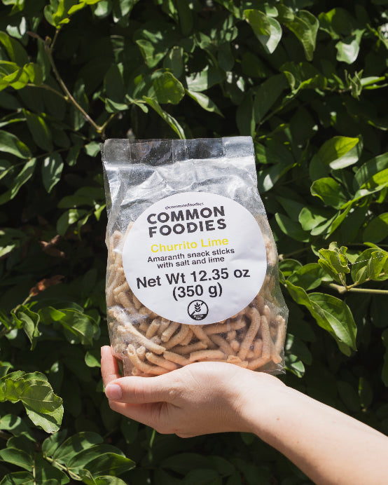 Hand holding a package of Common Foodies amaranth snack sticks against a green leafy background